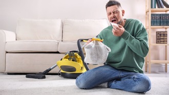 Aspirapolvere e uomo seduto sul tappeto che starnutisce